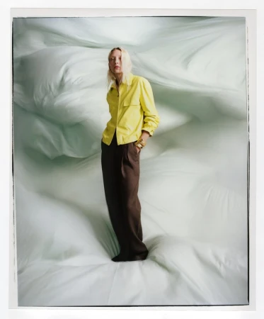 A stylish adult female poses in a yellow shirt and brown pants against a light-green background, full-length in a fashion-style photograph.