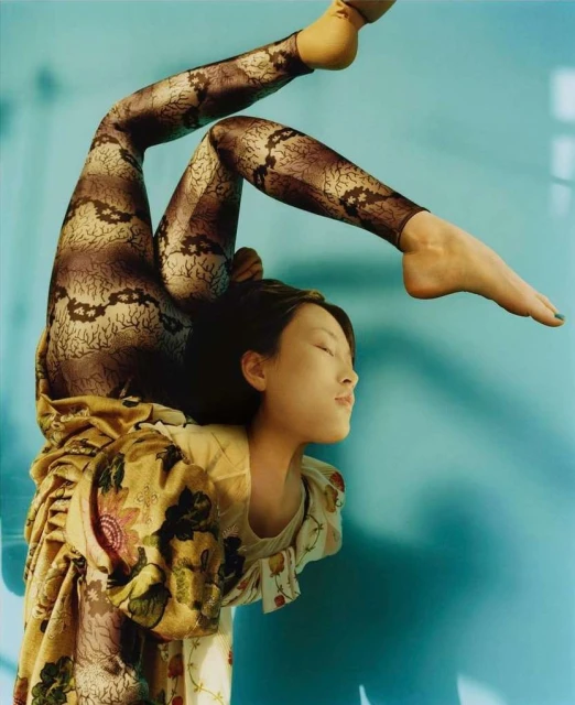 A striking artistic fashion shot features a person bending over with patterned leggings, a floral dress, and a blue seamless background.
