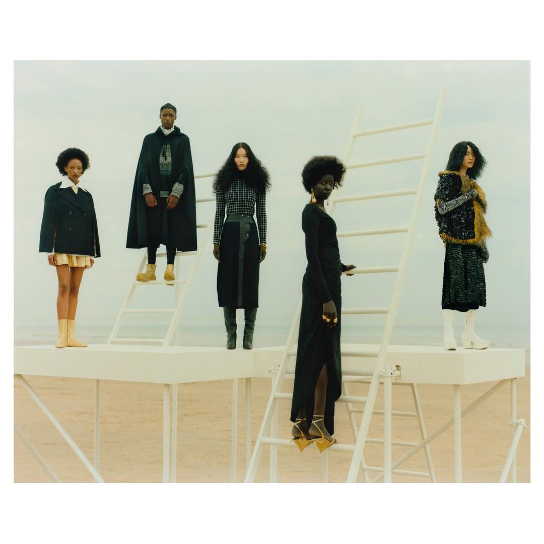 Five fashion models stand on white platforms in a landscape scene, wearing stylish clothing including long skirts, capes, and dresses.