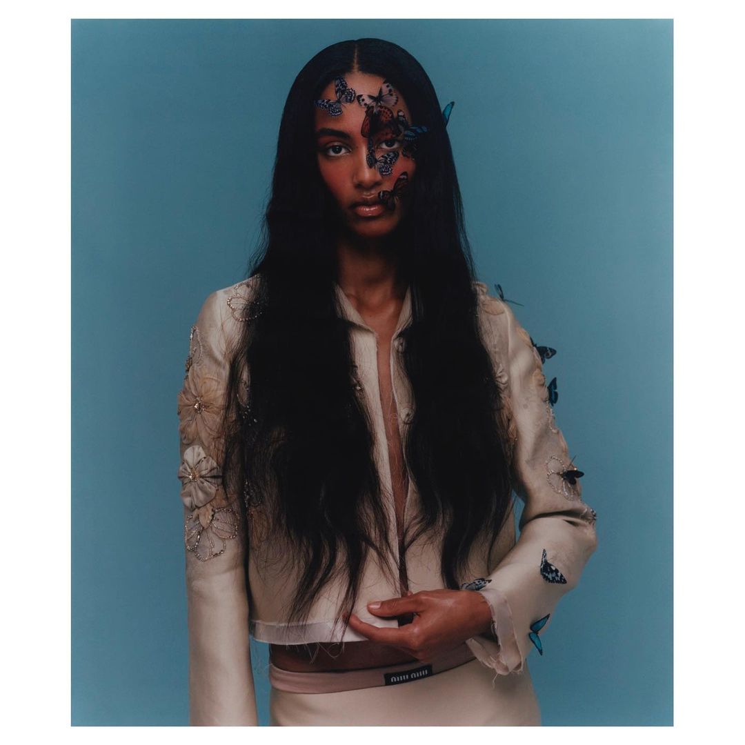 A model with long dark hair, adorned with butterfly accents and wearing a light-colored jacket, poses against a solid blue backdrop.
