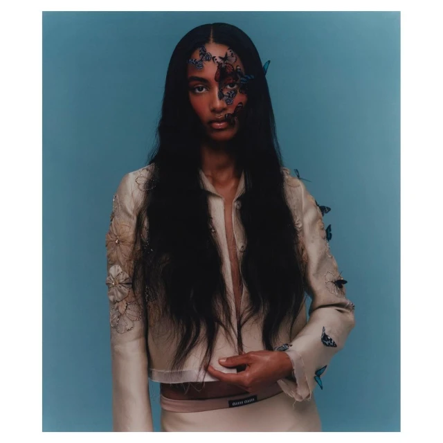 A model with long dark hair, adorned with butterfly accents and wearing a light-colored jacket, poses against a solid blue backdrop.