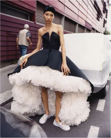 Model in strapless black gown with voluminous white ruffled underskirt standing on street by car