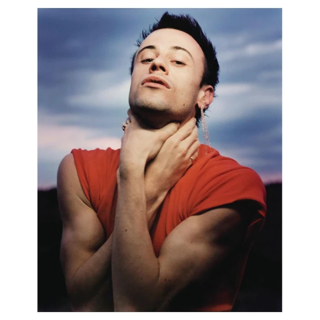 Man in a red shirt, hands at his neck, poses against a blue sky with pink clouds; earring glitters. Outdoors, portrait orientation, eye-level angle.