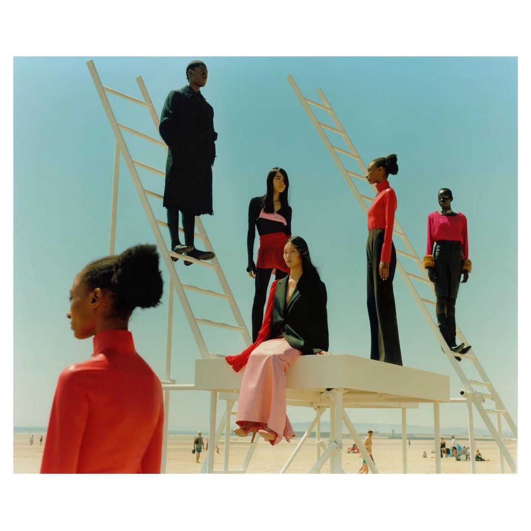 Fashion editorial photo of six models in red and black clothing, posing on a white platform and ladders, set on a sunny beach.