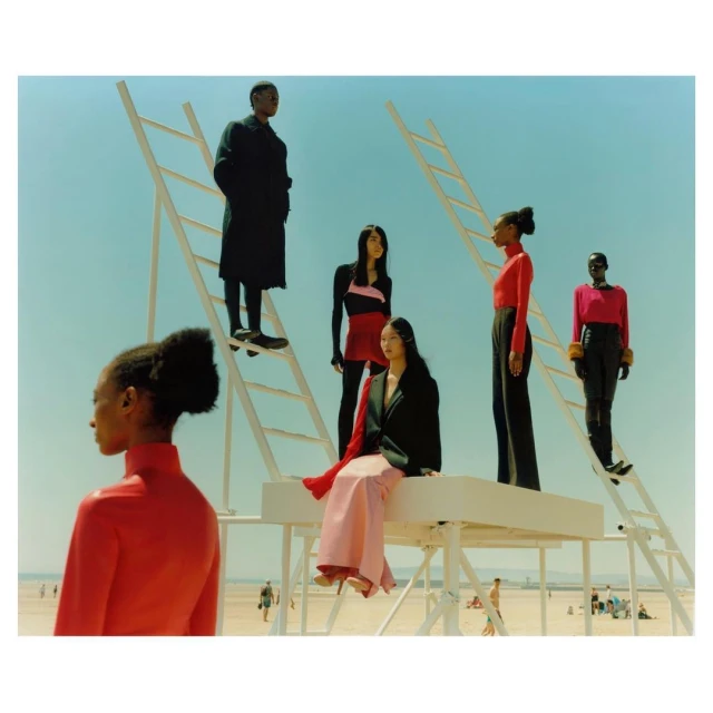 Fashion editorial photo of six models in red and black clothing, posing on a white platform and ladders, set on a sunny beach.