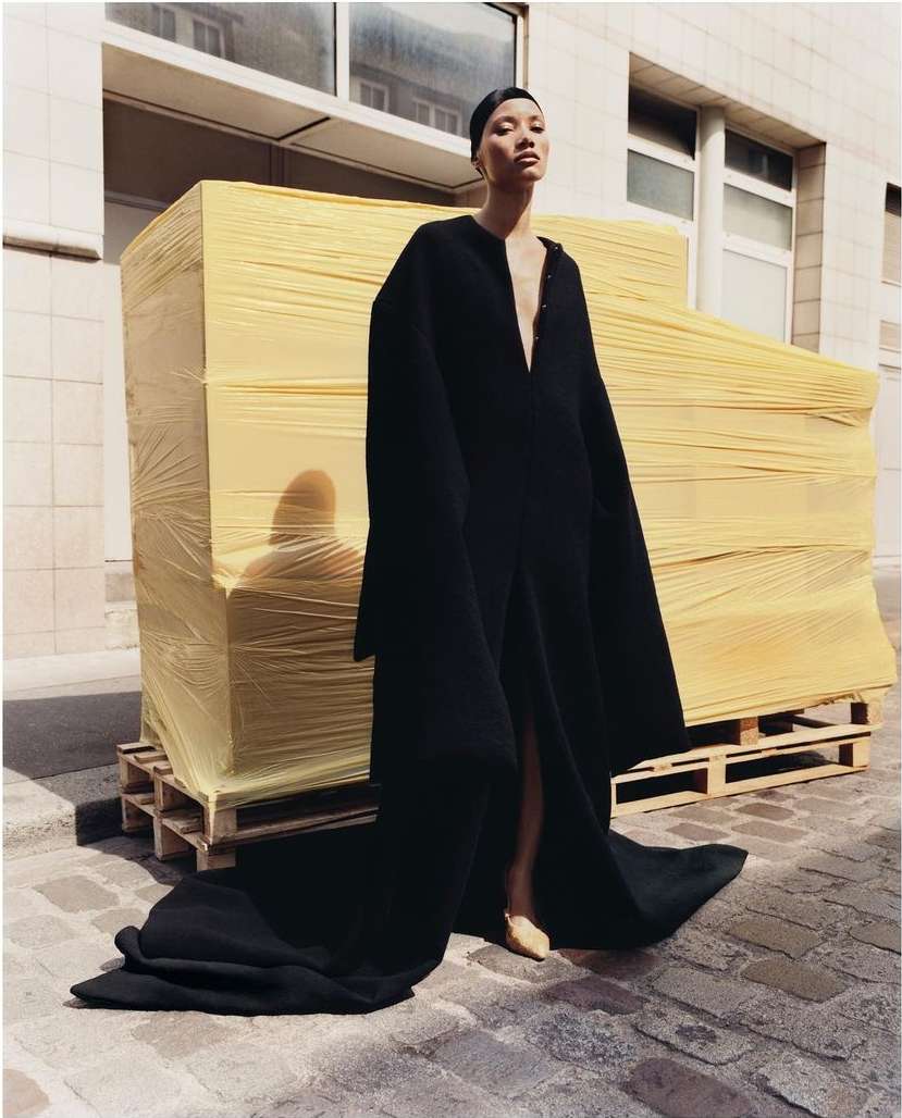 An Asian female fashion model poses outdoors in a long black coat. Wrapped construction materials sit behind her on a wood pallet.
