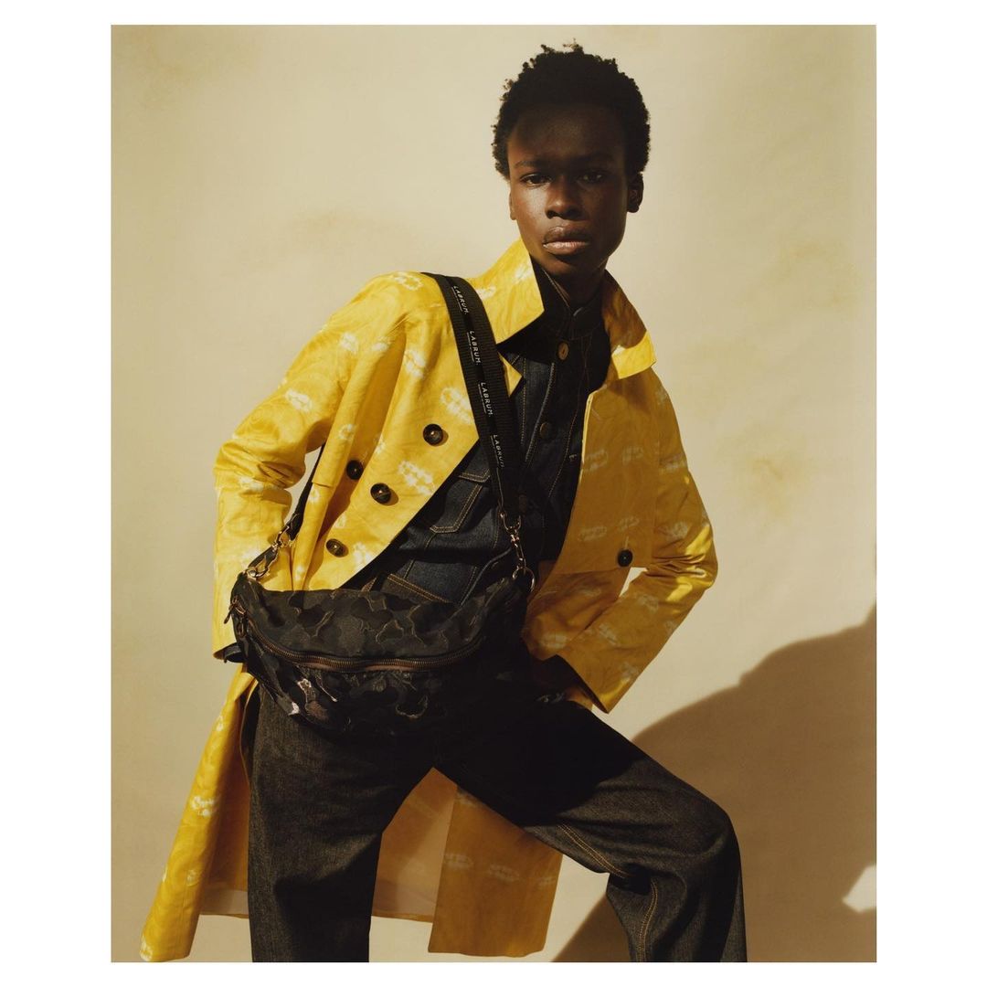 A Black fashion model in a yellow patterned coat, dark denim outfit, and a cross-body bag poses indoors against a neutral backdrop.
