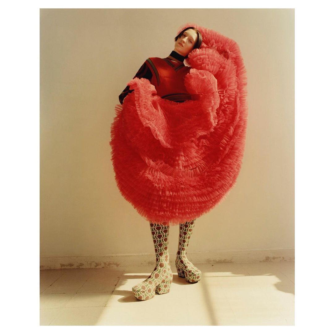 Fashionable adult female stands indoors, wearing a layered red tulle dress, dark turtleneck, patterned tights, and high platform boots.