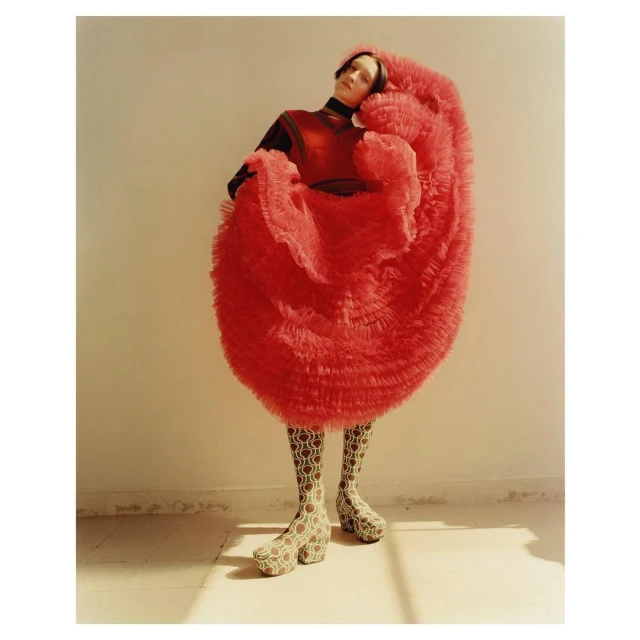 Fashionable adult female stands indoors, wearing a layered red tulle dress, dark turtleneck, patterned tights, and high platform boots.