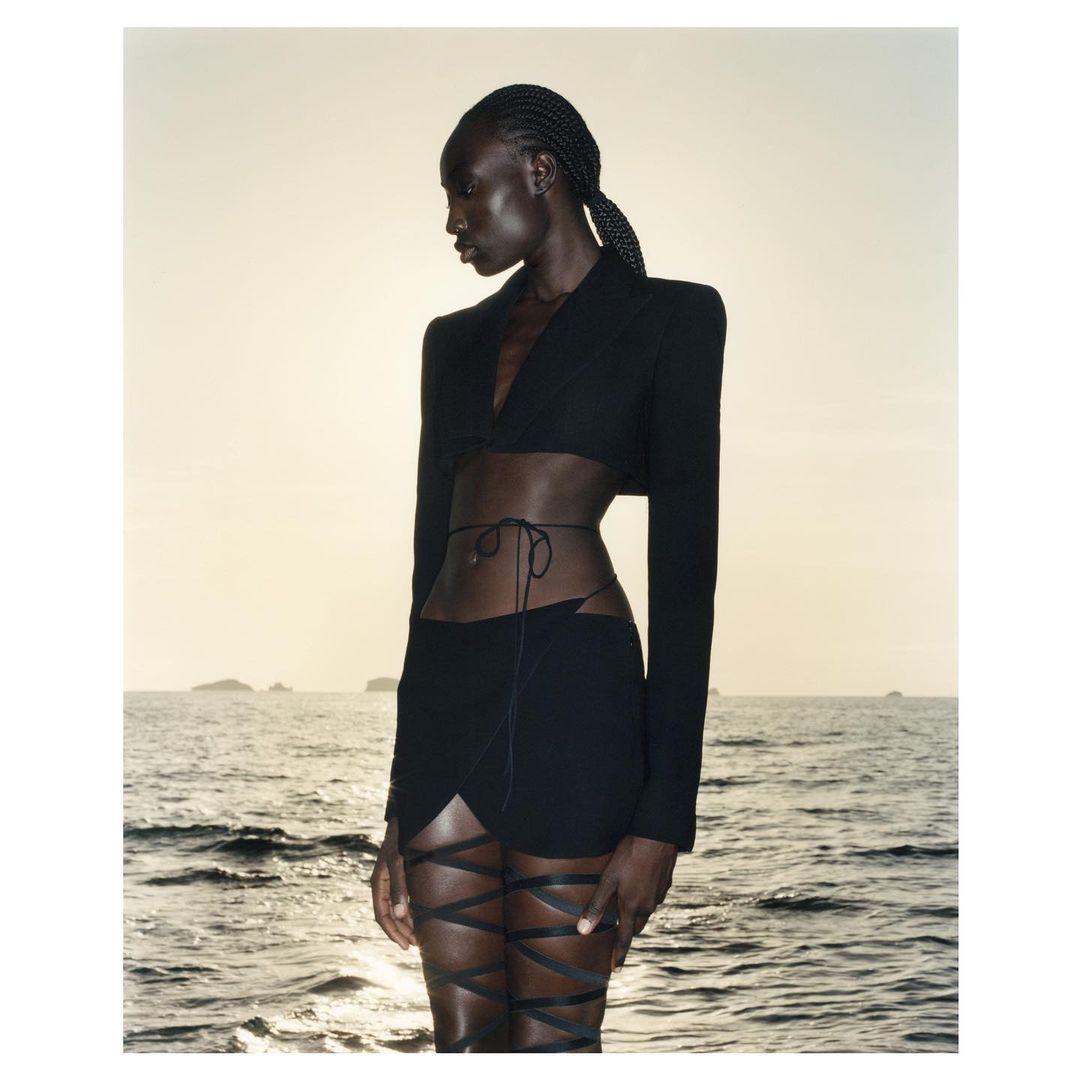 Black model with braided hair wearing a black crop top and skirt with leg ties standing by the ocean at sunset.