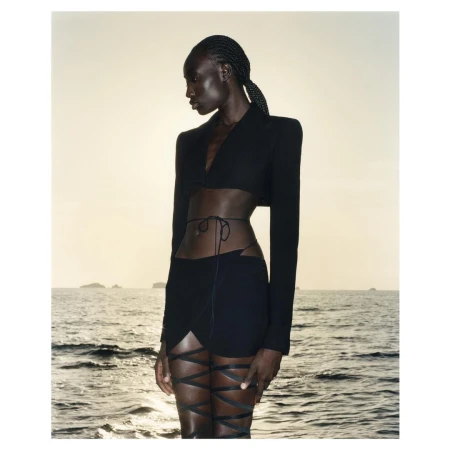 Black model with braided hair wearing a black crop top and skirt with leg ties standing by the ocean at sunset.