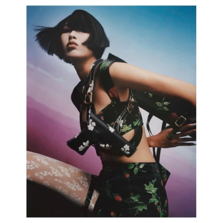 Model with dark bob hairstyle wearing a floral print outfit and carrying matching bags against a gradient sky.