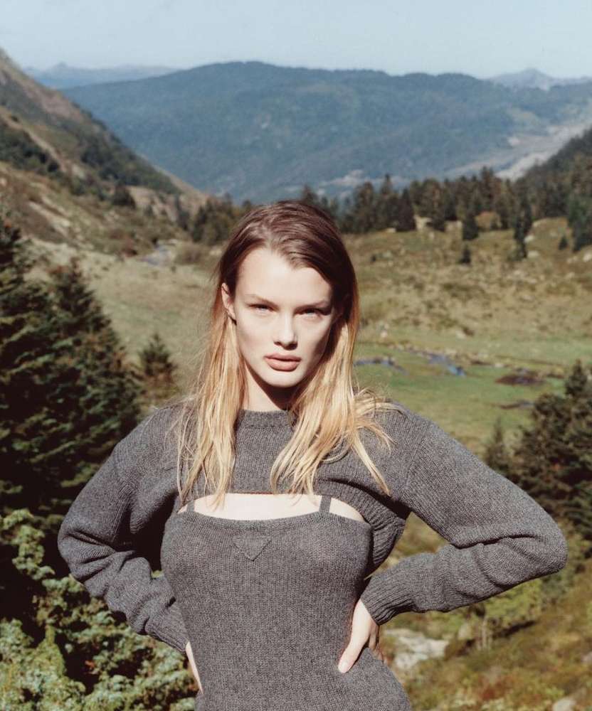 A blond woman poses in a grey knit dress and shrug with a mountain range and trees visible in the background, outdoor fashion shoot.