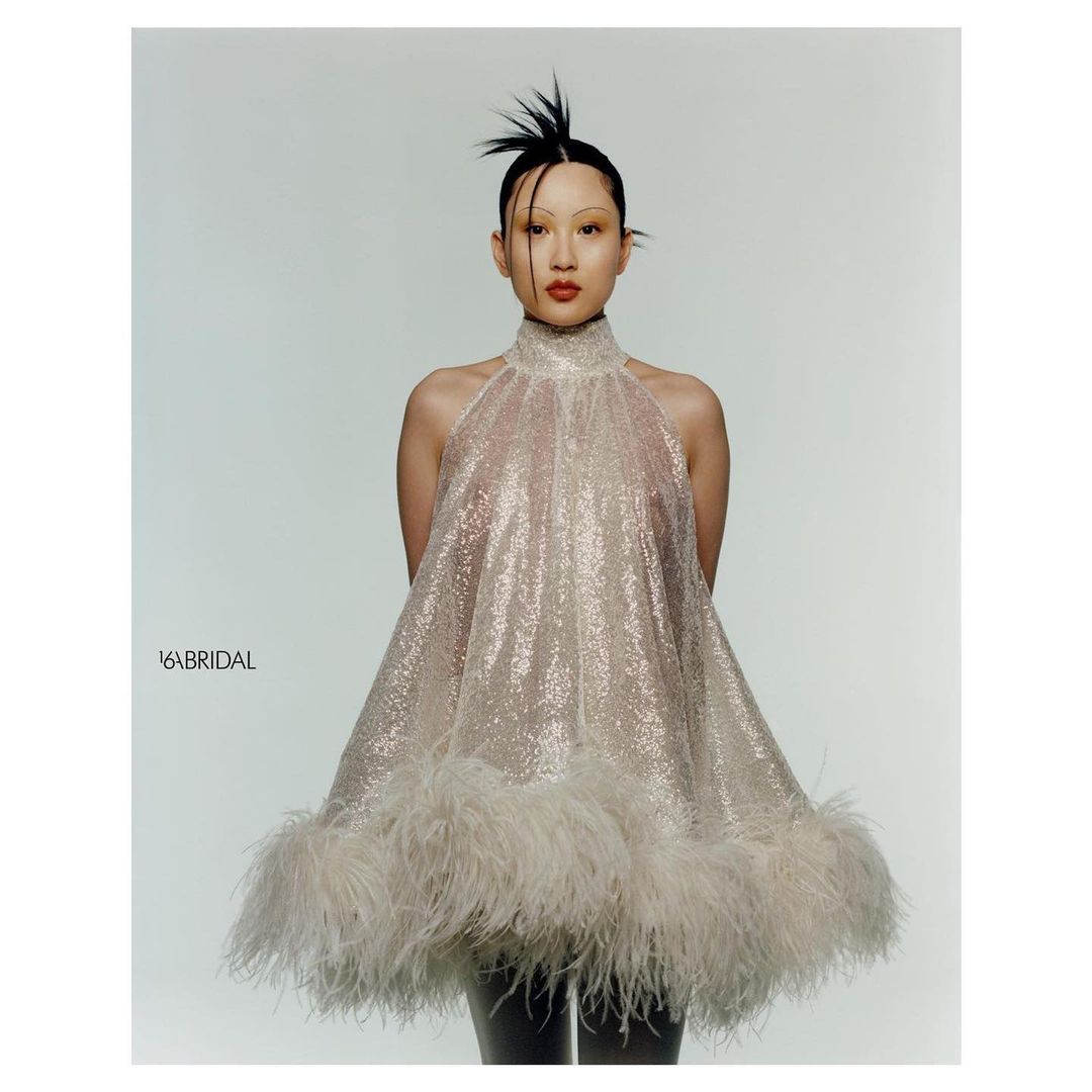 An adult female poses in a light studio wearing a short, sparkling halter dress with feather trim, hair styled up in spikes, red lipstick.