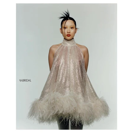 An adult female poses in a light studio wearing a short, sparkling halter dress with feather trim, hair styled up in spikes, red lipstick.