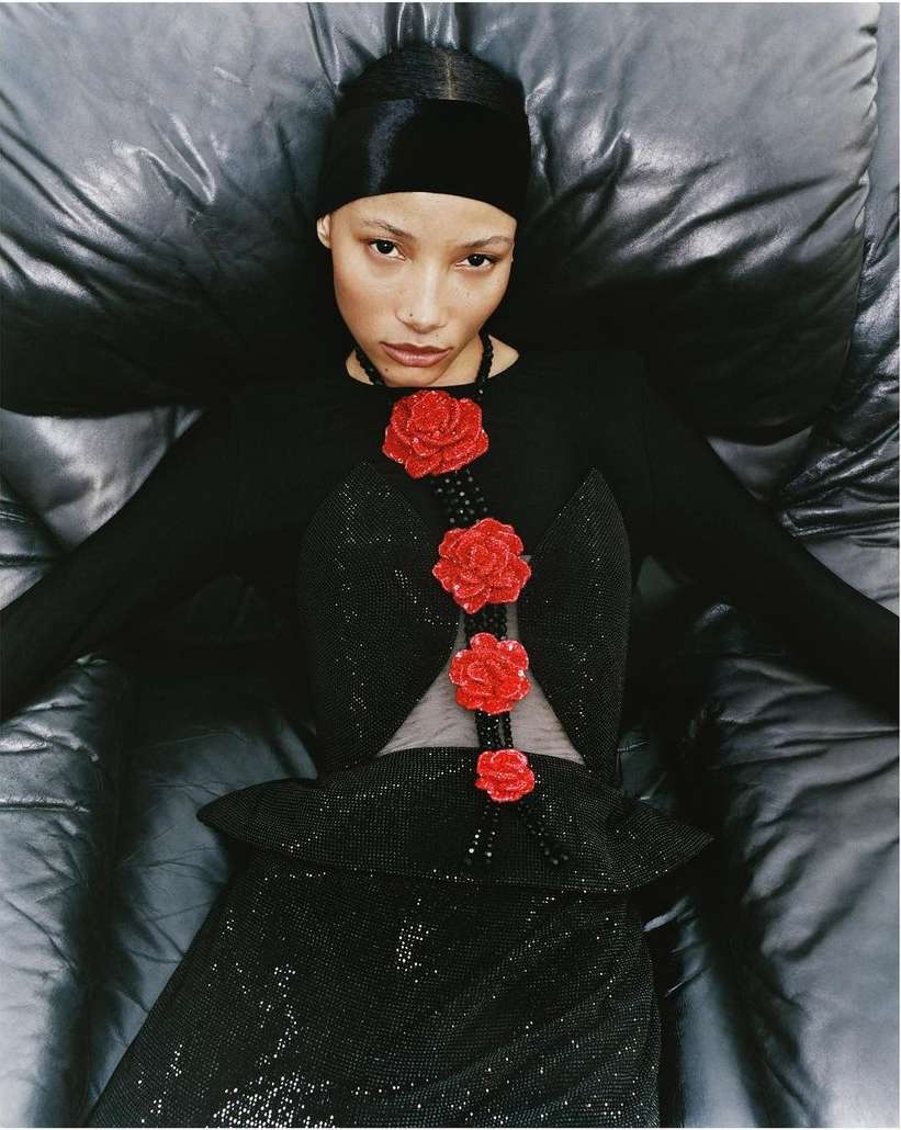 Woman in elegant black dress with red rose embellishments lies back on a glossy black surface, viewed from above, in a fashion-forward pose.