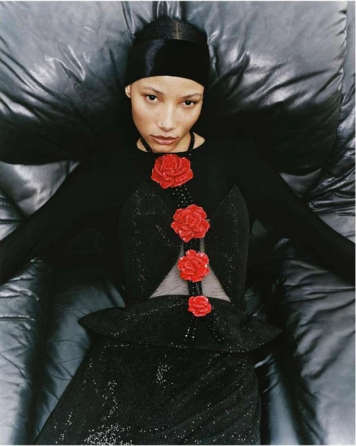 Woman in elegant black dress with red rose embellishments lies back on a glossy black surface, viewed from above, in a fashion-forward pose.