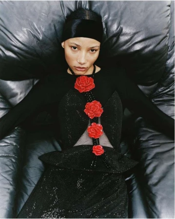 Woman in elegant black dress with red rose embellishments lies back on a glossy black surface, viewed from above, in a fashion-forward pose.