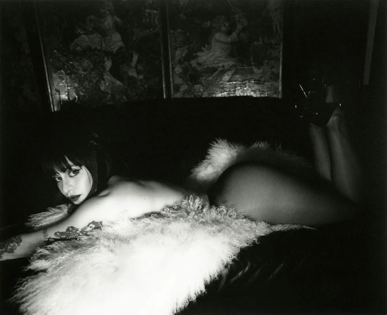 Monochrome portrait of a reclining woman with dark hair and tattoos, lying on a dark surface with a white fluffy throw, indoor setting.
