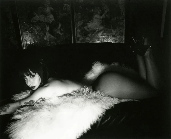 Monochrome portrait of a reclining woman with dark hair and tattoos, lying on a dark surface with a white fluffy throw, indoor setting.