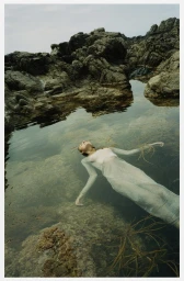 Woman in a sheer dress floating on her back in a calm rocky tidal pool filled with seaweed