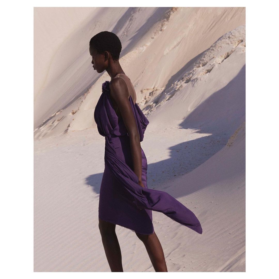 Elegant Black woman in a purple dress walking in a bright desert landscape. Fashion editorial image with visible light and shadows.