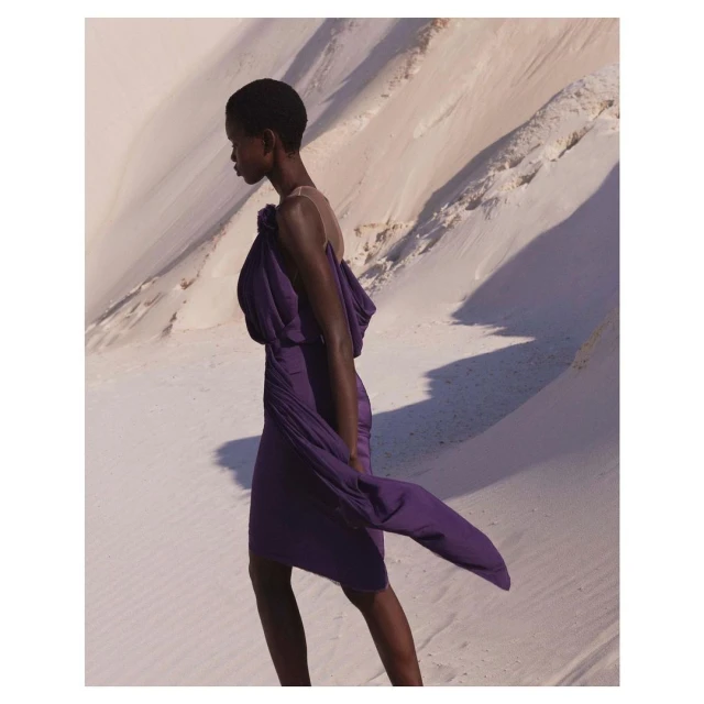 Elegant Black woman in a purple dress walking in a bright desert landscape. Fashion editorial image with visible light and shadows.