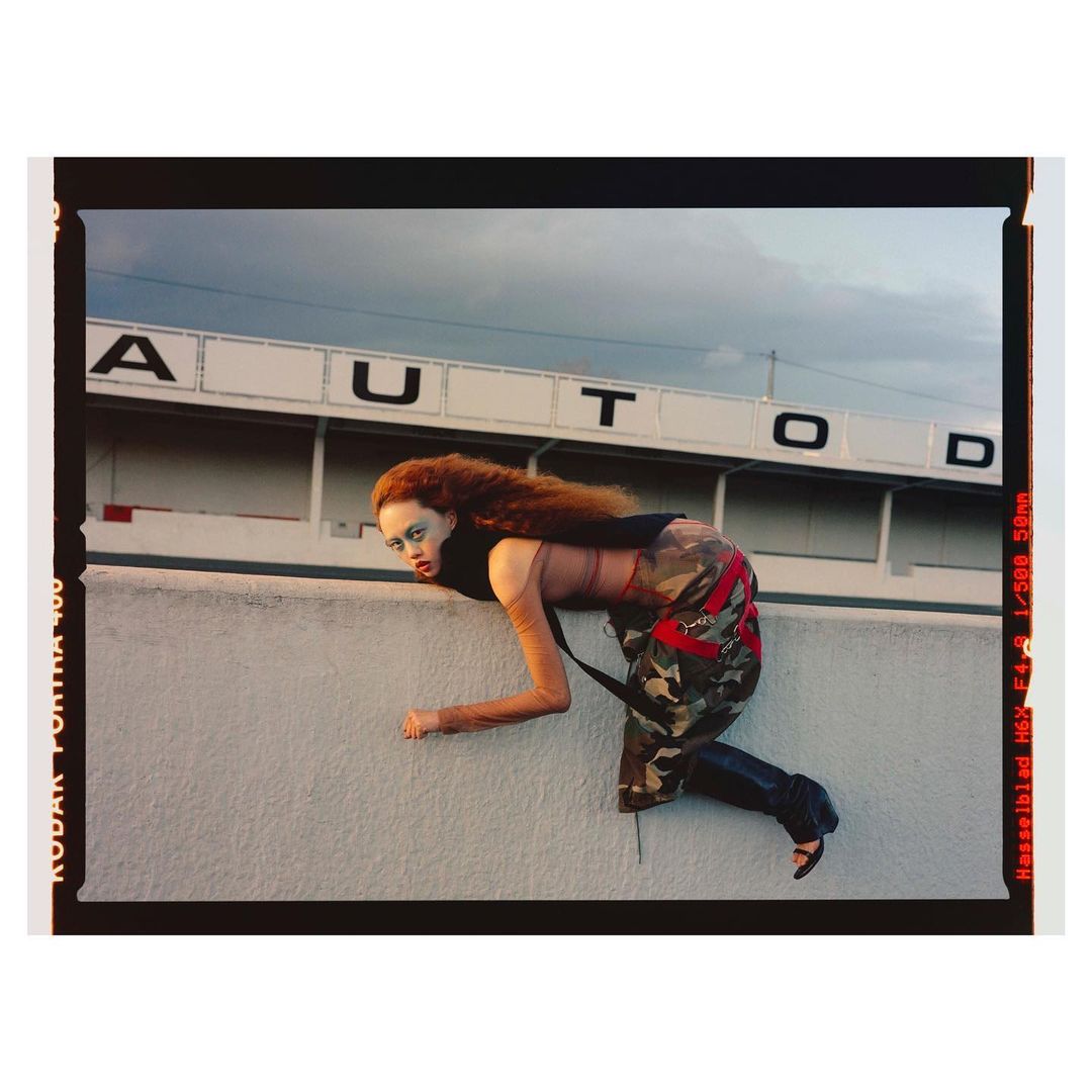 Fashion photography of a woman with red hair, wearing camouflage pants, black top and heels, crawling along a white wall outdoors.