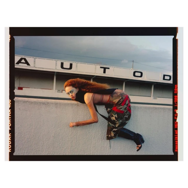 Fashion photography of a woman with red hair, wearing camouflage pants, black top and heels, crawling along a white wall outdoors.