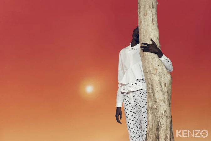 Model wearing a white shirt and patterned trousers embracing a tree trunk against a gradient orange sky with sun