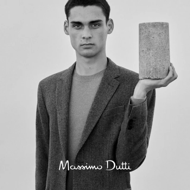 Monochrome portrait of a male model in a gray blazer holding a concrete cylinder, posing against a white background, featuring the Massimo Dutti logo.