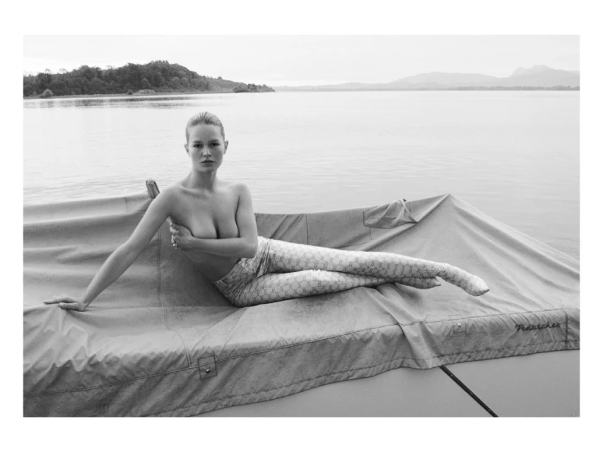 Monochrome portrait of a fair-skinned adult woman with mermaid-style leggings, reclining on a covered structure with a lake backdrop.