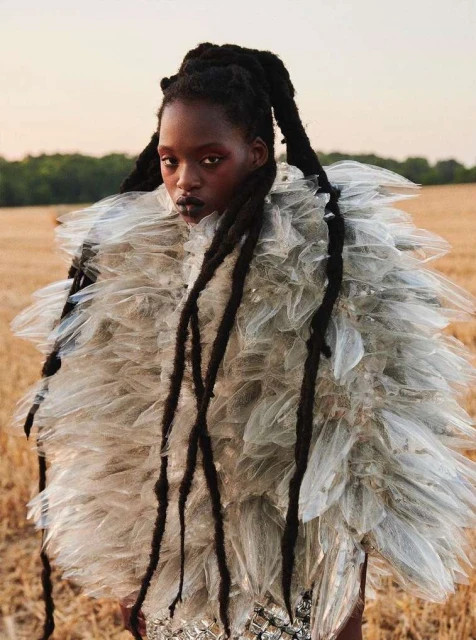 Fashionable woman with dark skin and long dreadlocks wearing a feathered dress in an outdoor field during daytime.