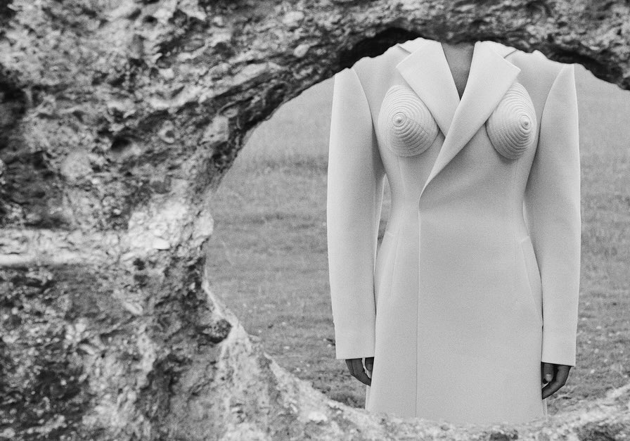 Monochrome image of a headless figure in a sculpted white coat, standing outdoors framed by a rock formation with a grassy background.
