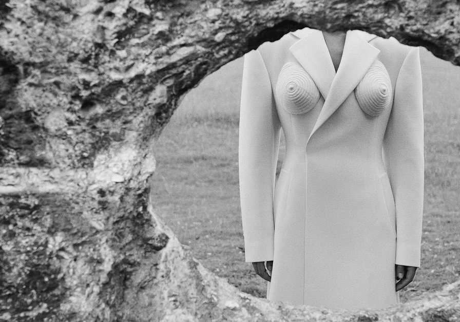 Monochrome image of a headless figure in a sculpted white coat, standing outdoors framed by a rock formation with a grassy background.
