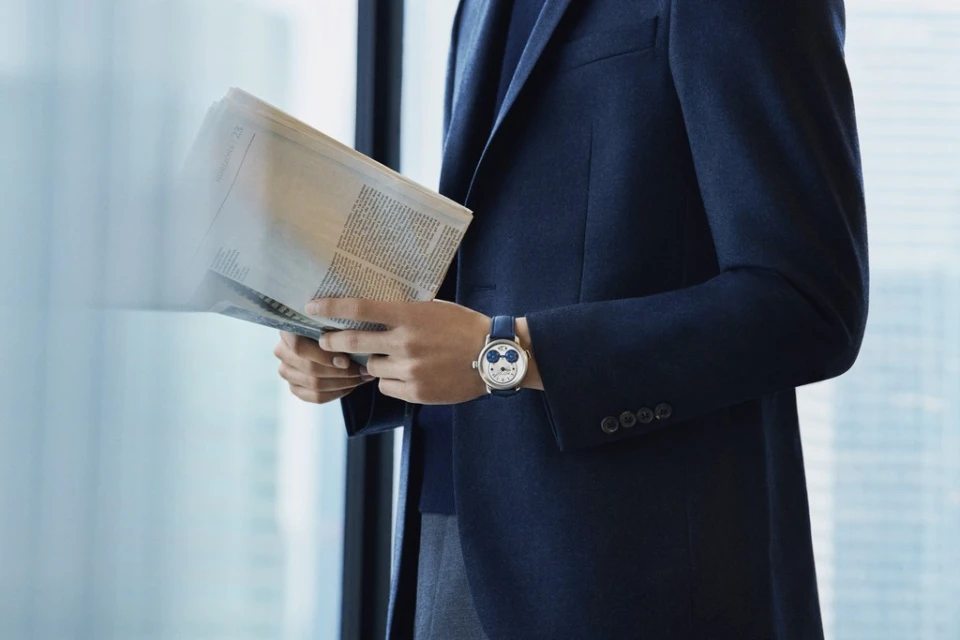 Man in a navy suit reads a newspaper near a window with a watch on his wrist. The watch has a cartoon-character design.