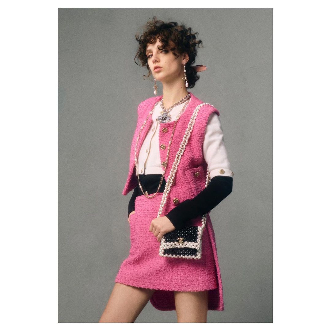 Woman in pink tweed skirt suit with pearl trim and black and white bag poses in front of a neutral gray background in studio shot.