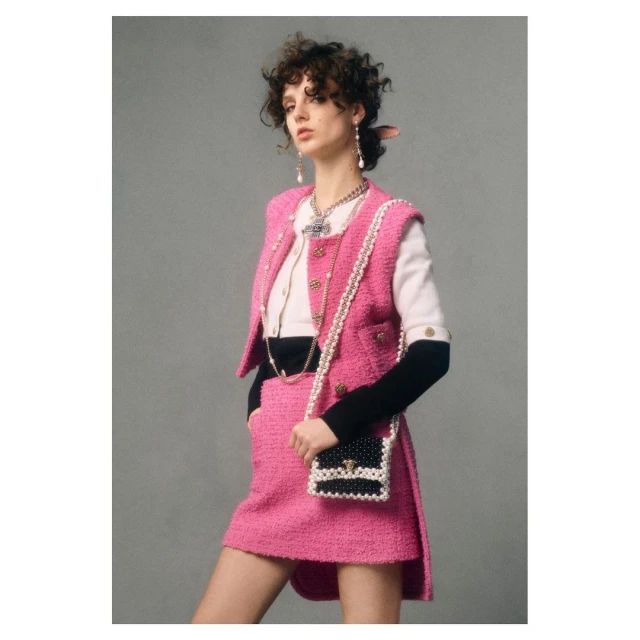 Woman in pink tweed skirt suit with pearl trim and black and white bag poses in front of a neutral gray background in studio shot.