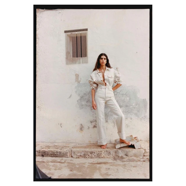 Woman in light denim co-ord poses outdoors near weathered wall with a small window; bare feet, long brown hair, full-length shot.