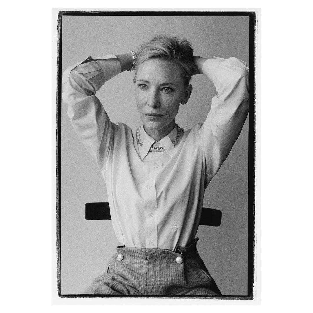 Black and white portrait of a woman in a white button-down shirt and patterned pants, sitting on a black chair with her hands behind her head.