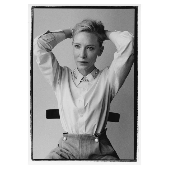 Black and white portrait of a woman in a white button-down shirt and patterned pants, sitting on a black chair with her hands behind her head.