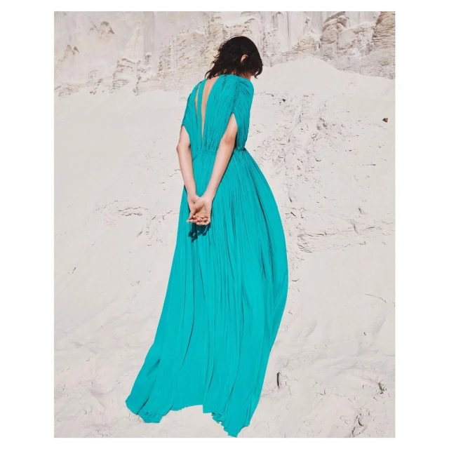Back view of woman in a long turquoise dress on a sandy hill. Fashion shot with natural light showing fabric texture and desert landscape.