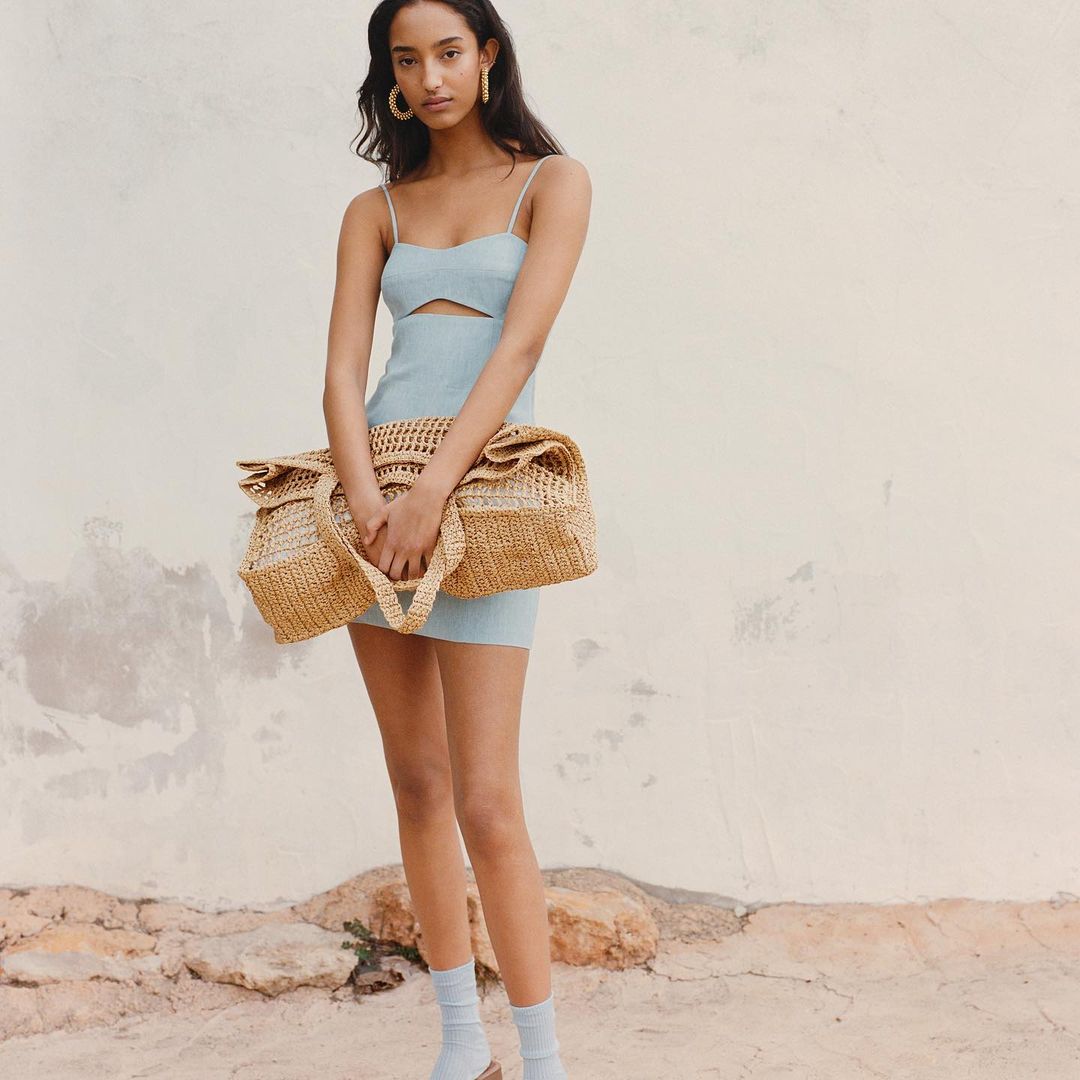 Portrait of an adult woman in a light blue dress holding a tan woven bag in an outdoor setting with a textured wall background.