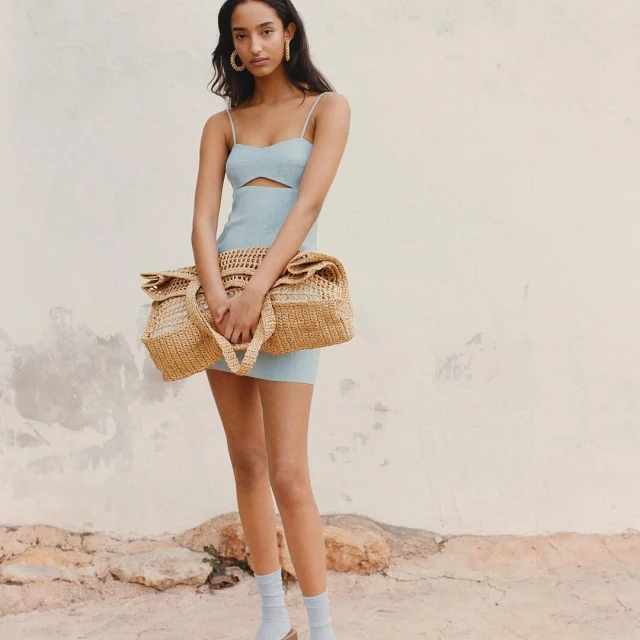 Portrait of an adult woman in a light blue dress holding a tan woven bag in an outdoor setting with a textured wall background.