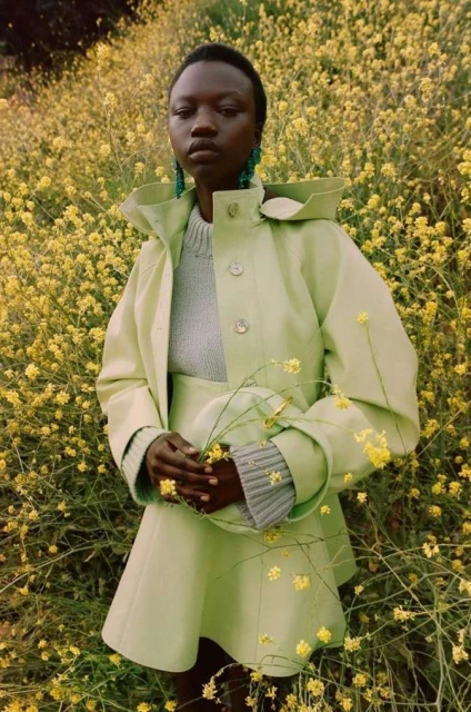Portrait of a stylish Black woman in a matching green jacket and skirt, holding wildflowers in a yellow field, wearing earrings.
