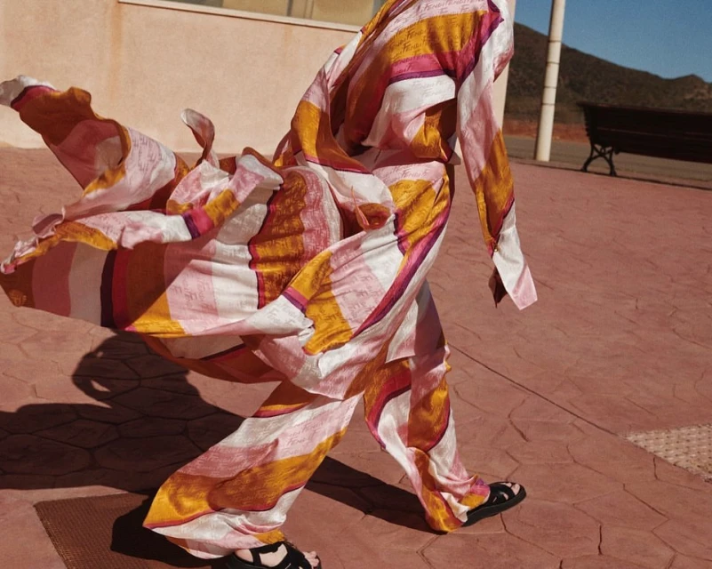 Person in pink and orange striped silk pajamas walking on a terracotta tiled patio near bench with a mountain in the background.