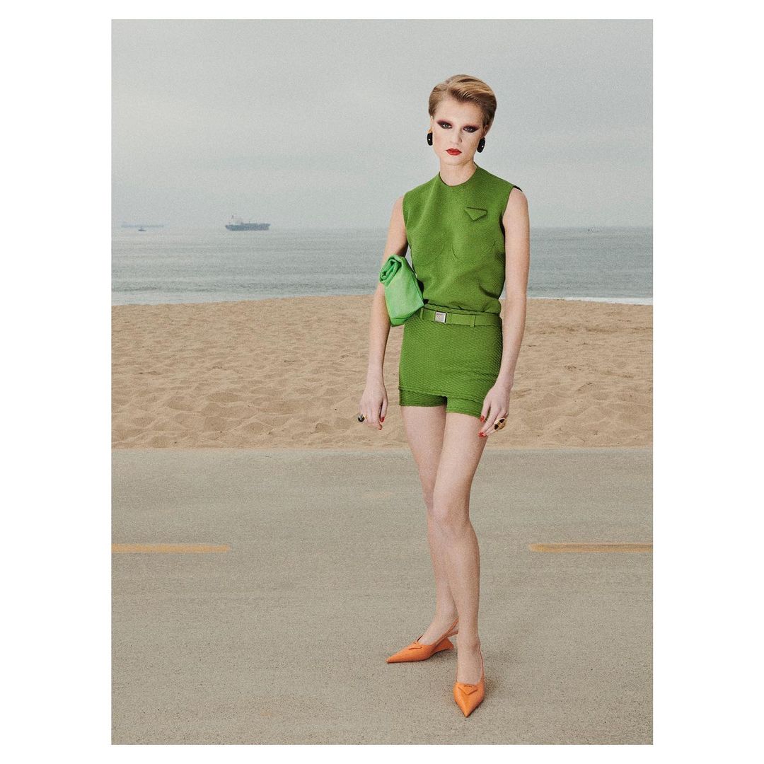 An adult female fashion model poses outdoors on pavement in a short green outfit with a matching bag and orange shoes, with the sea in the background.