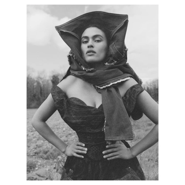 Black and white portrait of a woman in a denim dress and head covering in a rural setting with a cloudy sky and trees in background.