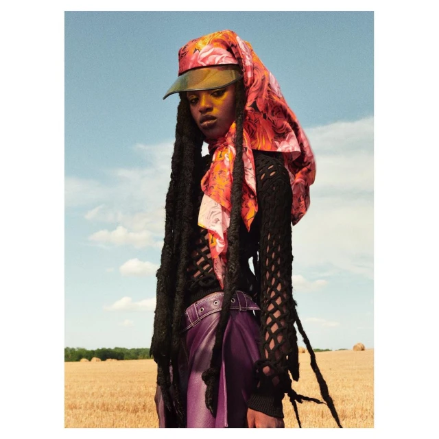 Woman with long dark braids poses in a farm field wearing a floral hat, black sweater, and purple skirt against a blue sky.