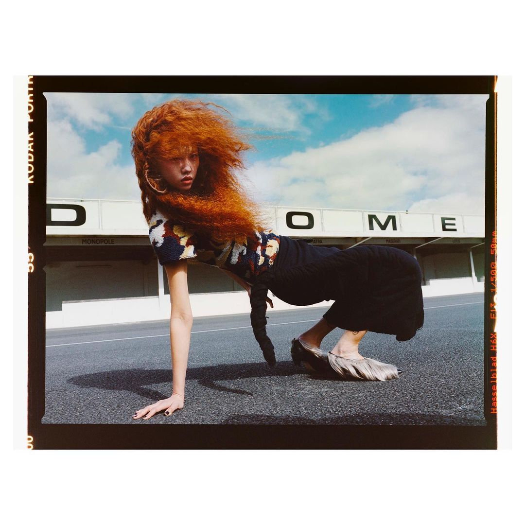 Fashion shot of a red-haired woman in black and patterned clothing posing on pavement outside a building under a cloudy sky.
